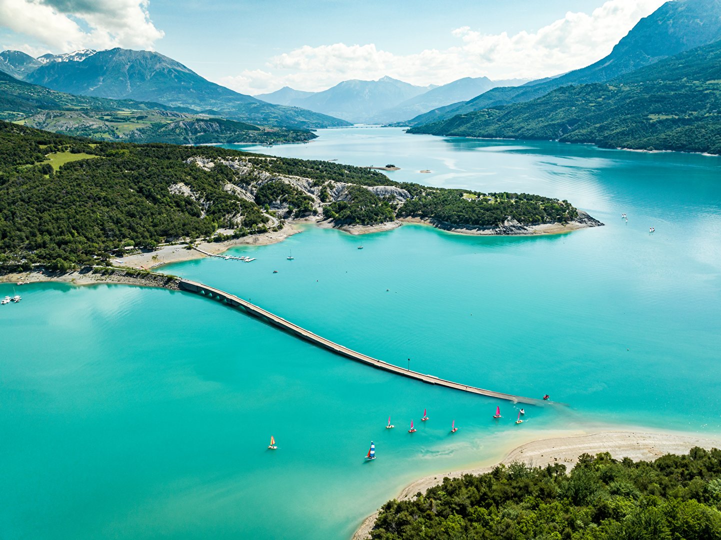 Vue aérienne du lac de Serre-Ponçon dans les Hautes-Alpes : le pont de Savines-le-Lac traverse l'eau turquoise, des voiliers naviguent, les montagnes en toile de fond